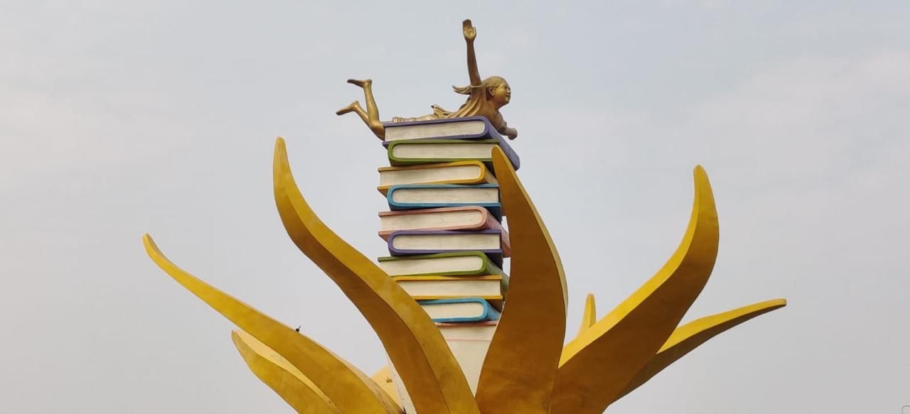Detail: a golden figure lying on a stack of coloured books, arm raised, framed by leaf forms