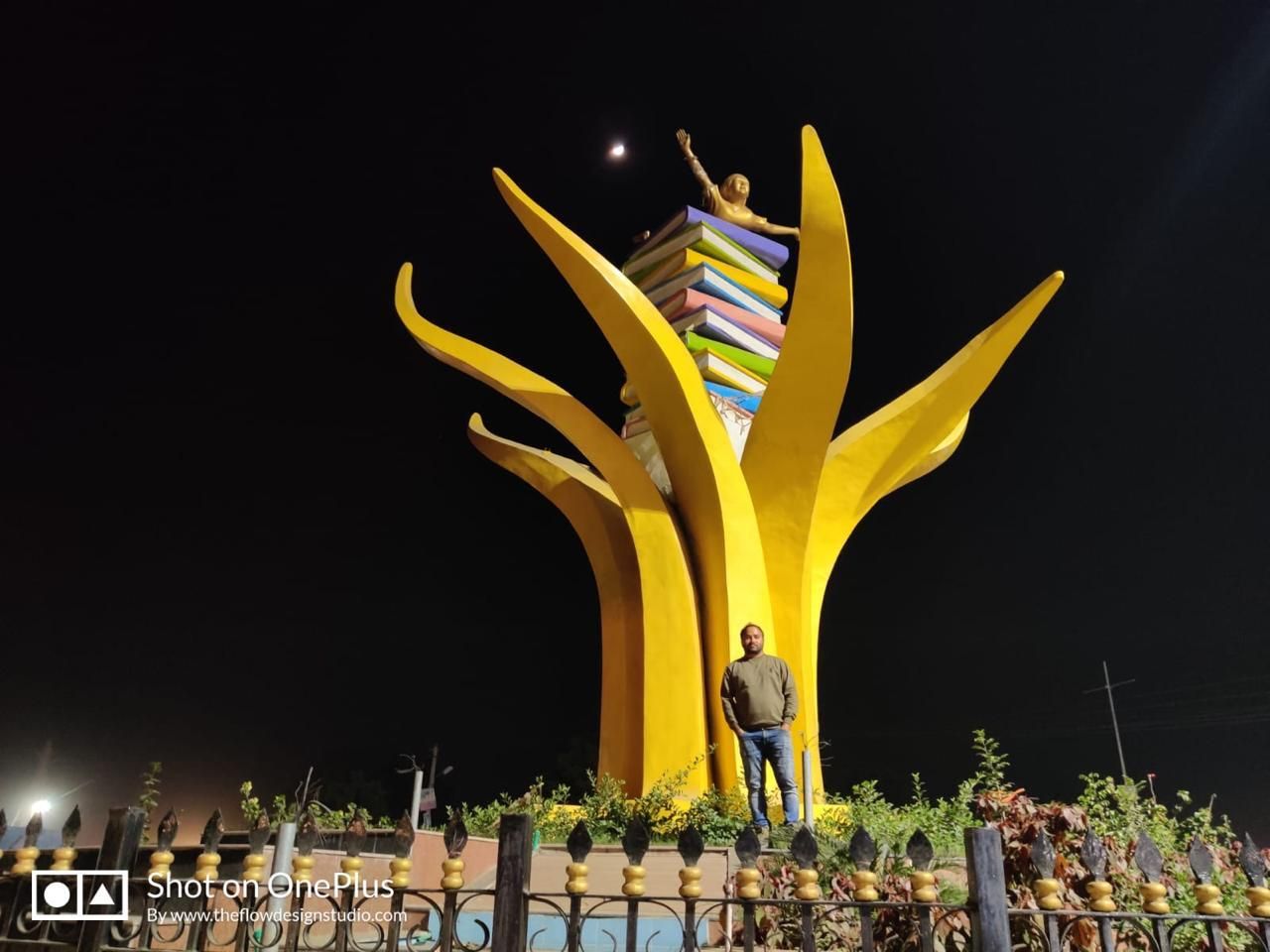 The book sculpture lit at night, with Pawan Kumar Bhatt standing at its base