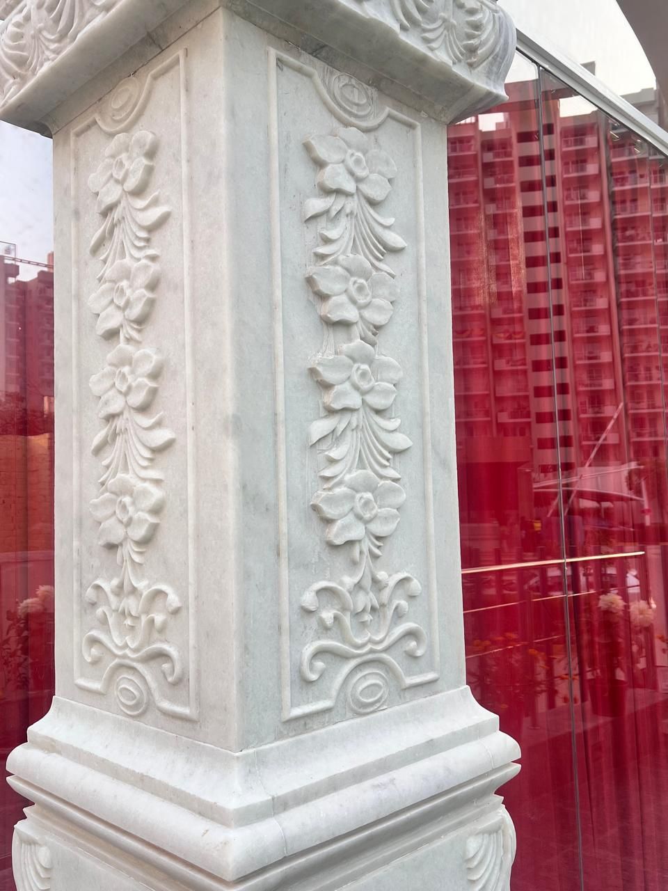 A marble pillar carved with climbing floral panels, red curtains reflected in the glass behind