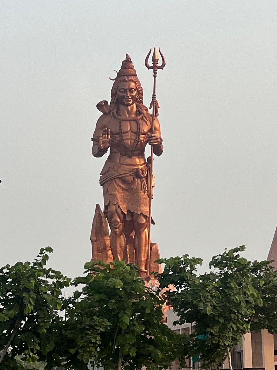 Copper-toned standing Lord Shiva sculpture with trident rising above trees, Panipat