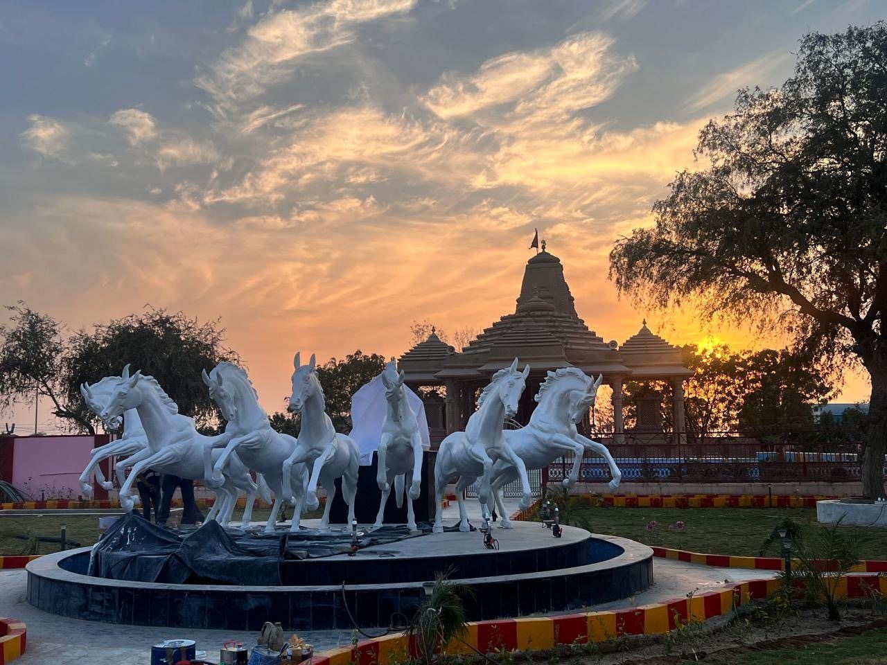Seven white rearing horses on a round fountain plinth at sunset, with a sandstone temple behind