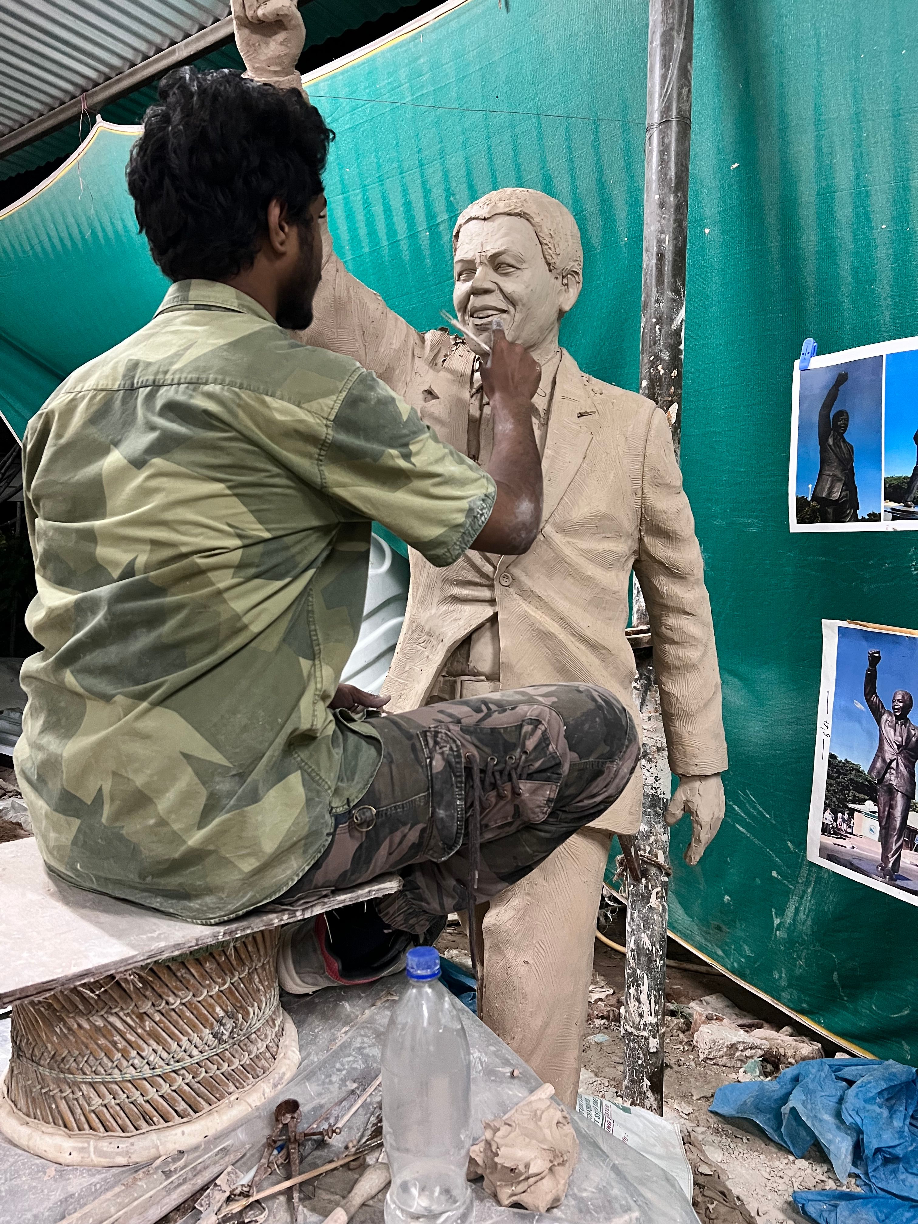 Sculptor seated on a stool, working the face of a full-size clay portrait figure, with reference photographs pinned beside it