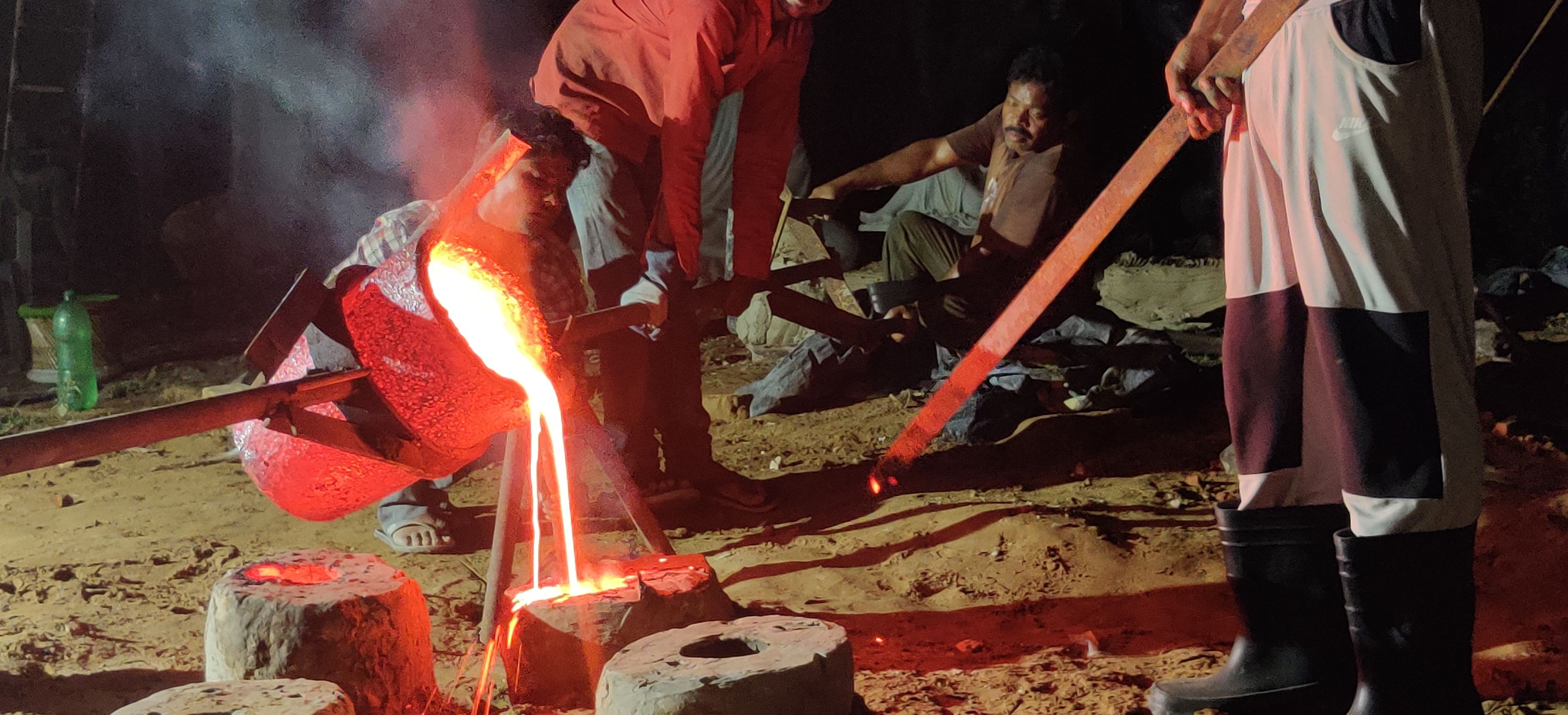 Foundry workers at night tilting a glowing crucible, pouring molten metal into a row of moulds set in sand