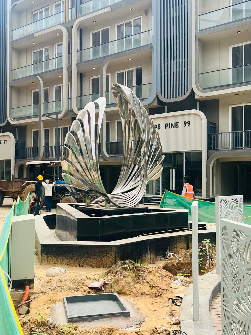 A mirror-polished stainless-steel wing sculpture on a fountain plinth, in a residential courtyard under construction
